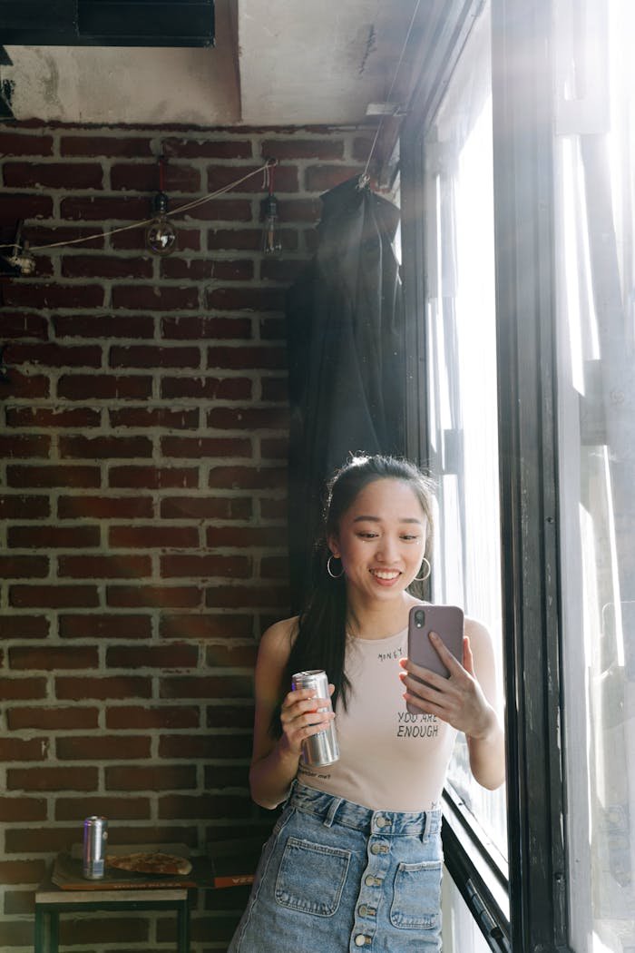 Asian woman enjoys a soda while taking a selfie in a brick-walled loft.